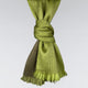 A green fabric scarf is tied into a stylish knot, with fringed ends hanging loosely, against a plain light gray background.