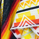 A woven fabric features bold geometric patterns with triangles and stripes in vibrant yellow, orange, red, and black. It is slightly folded, displaying textured, colorful designs in close-up detail.