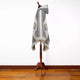 A mannequin displays a hooded poncho with geometric patterns and fringed edges. It stands in a plain-white room with a wooden floor, highlighting the textile's intricate design.