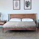 A wooden bed with white pillows and a patterned bedspread is centrally placed. On either side are wooden nightstands, and two framed leaf prints hang on the wall above.