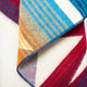 A colorful, folded wool blanket displays a pattern of stripes in blue, orange, and red hues against a soft white background, creating a cozy and vibrant texture.