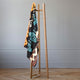 A wooden ladder-style rack holds a colorful patterned blanket with geometric designs, set in a minimalist room with a light gray wall and parquet flooring.