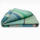 Sucua baby alpaca wool blanket – a folded blanket rests on a flat, plain surface, featuring green, blue, and beige horizontal stripes with a soft texture