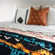 Pillows with geometric patterns rest on a bed, covered by a vibrantly patterned blanket featuring red, blue, and orange designs. The room looks bright and cozy.