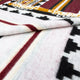 A woven blanket featuring geometric patterns in red, black, and mustard lines and shapes, along with a folded fabric corner, rests against a contrasting lighter background surface.