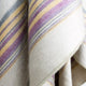 Quijos alpaca wool blanket – a wool blanket drapes gently, showcasing pastel stripes of purple, yellow, and gray, within a soft, textured fabric in a cozy indoor setting