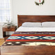 Qhusi extra large alpaca wool blanket – bed with a wooden frame and patterned blanket in geometric designs. Two pillows rest on the bed. A small plant sits on a wooden nightstand, with a wreath on the wall above