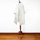 A white poncho drapes over a mannequin, positioned on a wooden floor against a plain white wall. The poncho features a simple, elegant design with a hood.