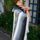 Palora lightweight alpaca wool hooded poncho – a person wrapped in a striped, fringed poncho stands with one hand near their face, in a garden setting with a blue-windowed building in the background