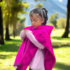 Child in a vibrant pink poncho looks down at hands in a lush, sunlit park with trees and mountains in the background.