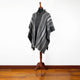 A dark gray poncho with white stripes is displayed on a wooden mannequin stand, set against a white wall and wooden floor.