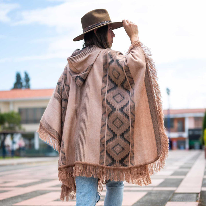 A person in a fringed poncho and hat stands outdoors, adjusting the hat brim. The background features a tiled pathway and distant buildings under a cloudy sky.