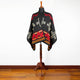 A mannequin displays a black poncho featuring bold red and geometric patterns, situated on a wooden stand against a white background, atop a wooden floor.