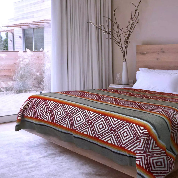 A bed with a geometric-patterned blanket and white pillows sits in a modern bedroom with a wooden headboard, large window, and a vase with branches on a nearby table.