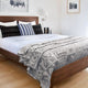 A wooden bed is covered with a white sheet, patterned pillows, and a gray geometric blanket. The room features wooden flooring, framed wall art, and soft lighting.