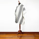 A cream and black patterned poncho is displayed on a wooden mannequin stand in a room with wood flooring and a white wall.