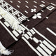 Kids Clint Eastwood alpaca wool poncho – a woven fabric features geometric patterns in cream on a rich brown background, adorned with a clover motif. Fringed edges add texture. The setting is flat and close-up