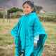 Cuyabeno kids alpaca hooded poncho – a young child stands smiling, wearing a bright turquoise poncho with geometric patterns, in a grassy outdoor area with blurred mountains and trees in the background