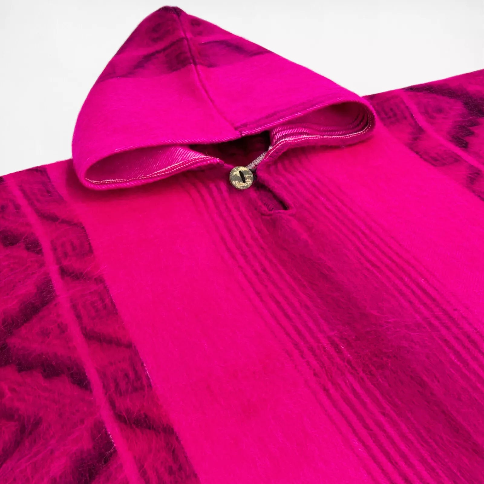 A bright pink hooded garment features geometric patterns and a wooden button. The fabric is textured and the hood is laid back, highlighting intricate designs in a soft material.