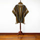 Poncho drapes on a wooden stand, featuring green, orange, and white geometric patterns. Fringed edges and rich colors provide contrast against a simple white wall and wooden floor.