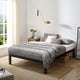 A neatly made bed with gray patterned bedding and pillows sits in a room with a white brick wall. There's a wooden nightstand holding books and a vase, under a gold wall lamp.