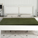 A bed with a green blanket rests against a white-paneled wall. It has two pillows, flanked by nightstands with lamps. A picture and plant decorate one nightstand.