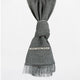 A gray scarf hangs with a knot at its center. It features an embroidered pattern strip and fringes at the bottom, set against a plain white background.