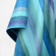 Pululahua baby alpaca wool blanket – a soft, multicolored fabric with diagonal blue, turquoise, and lavender stripes drapes gently. The colors blend smoothly, creating a harmonious and textured appearance against a plain background