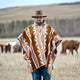 Man wears a patterned poncho and hat, standing in a field. Horses graze in the background, surrounded by rolling hills and trees, under a cloudy sky.
