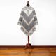 Poncho displayed on a wooden stand, featuring a geometric pattern in gray and beige tones, with buttons at the collar, set against a plain white background on a wooden floor.