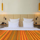 Putumayo baby alpaca wool blanket – a bed with white pillows and an orange-striped blanket is flanked by bedside tables with lamps and beige padded headboard, under two hanging yellow pendant lights in a minimalist room