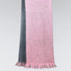 A two-toned scarf hangs vertically, blending soft pink and dark gray hues. The scarf features a textured knit pattern and fringe at the edges, set against a plain backdrop.