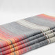 Ajilanga baby alpaca wool blanket – a neatly folded woolen blanket displays stripes in various shades of gray, red, orange, and cream. It rests against a simple white background