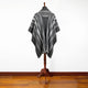 A striped, grey poncho is draped over a wooden mannequin stand on a polished wooden floor in a plain, white-walled room.