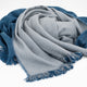 A soft, two-toned shawl with light gray and blue colors is draped elegantly, featuring fringed edges, laid against a plain white background.