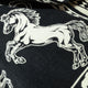 A white horse printed on black fabric, depicted in a rearing pose. The surrounding pattern includes geometric shapes, adding a dynamic and bold contrast to the image.