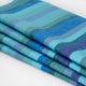 Pululahua baby alpaca wool blanket – a folded woolen blanket displays wide, horizontal blue and green stripes. It rests on a white surface, showcasing its soft, textured material and vibrant color pattern