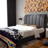 A bed with a dark, high-backed headboard is neatly made with white sheets and a colorful patterned blanket; it's in a modern bedroom with wooden wall decor and dark curtains.