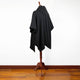A black hooded poncho is displayed on a wooden mannequin stand in a room with a polished wooden floor and a plain white wall.