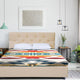 A bed with a tan, tufted headboard, covered in a colorful geometric blanket, stands in a modern bedroom with hanging lights, minimalist decor, and green plants.