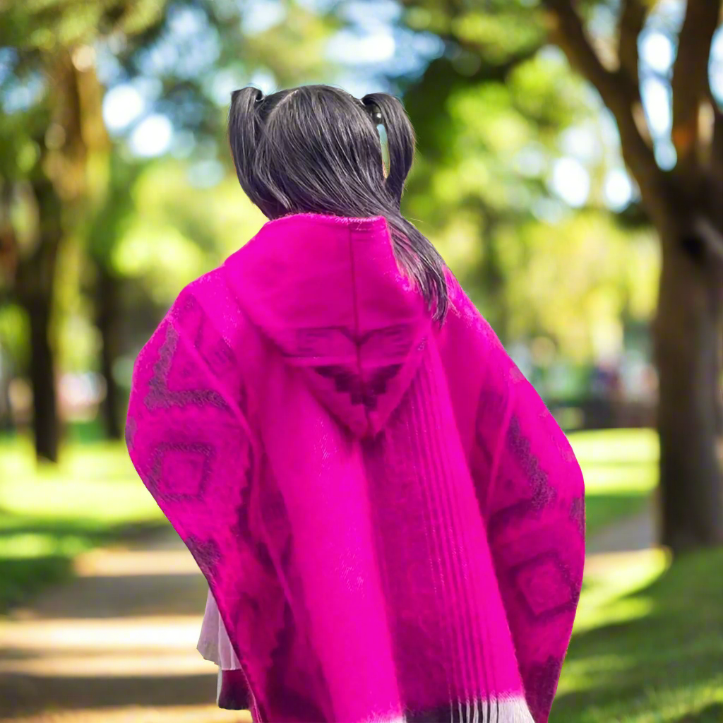 A person, with black hair in pigtails, wearing a bright pink poncho, walks through a tree-lined park path on a sunny day.