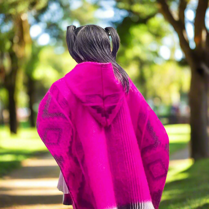 Macas kids alpaca hooded poncho – a person, with black hair in pigtails, wearing a bright pink poncho, walks through a tree-lined park path on a sunny day