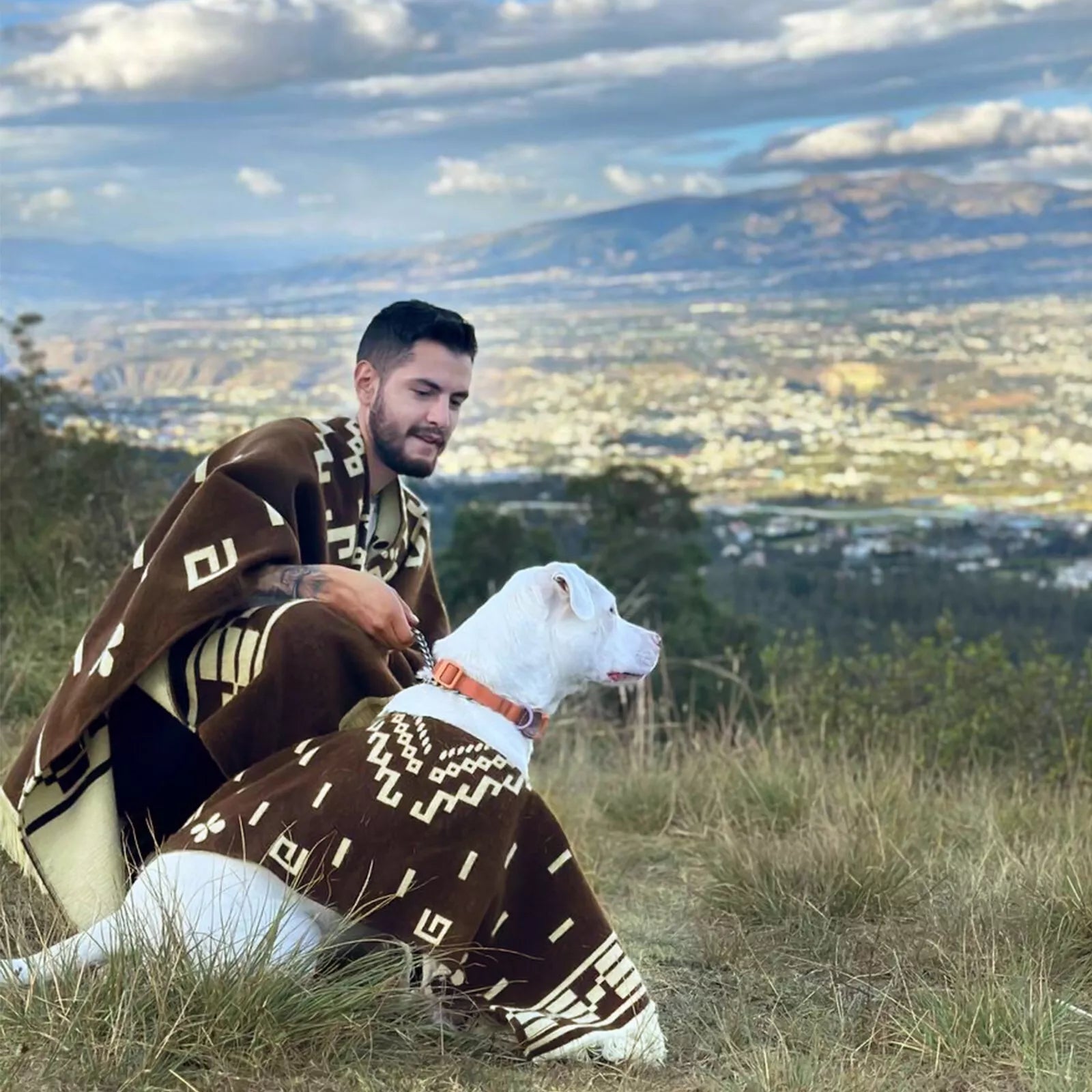 A man and a white dog, both wearing matching brown patterned ponchos, sit on grass overlooking a scenic valley and mountains under a cloudy sky.