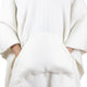 A person wears a soft, oversized white sweater with their hands tucked into the large front pocket, providing a sense of warmth and comfort in a minimalist setting.