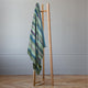 Imbabura baby alpaca wool blanket – a wooden clothes rack stands upright, holding a striped blanket with shades of green, blue, and purple. It is set against a plain gray wall and wooden floor