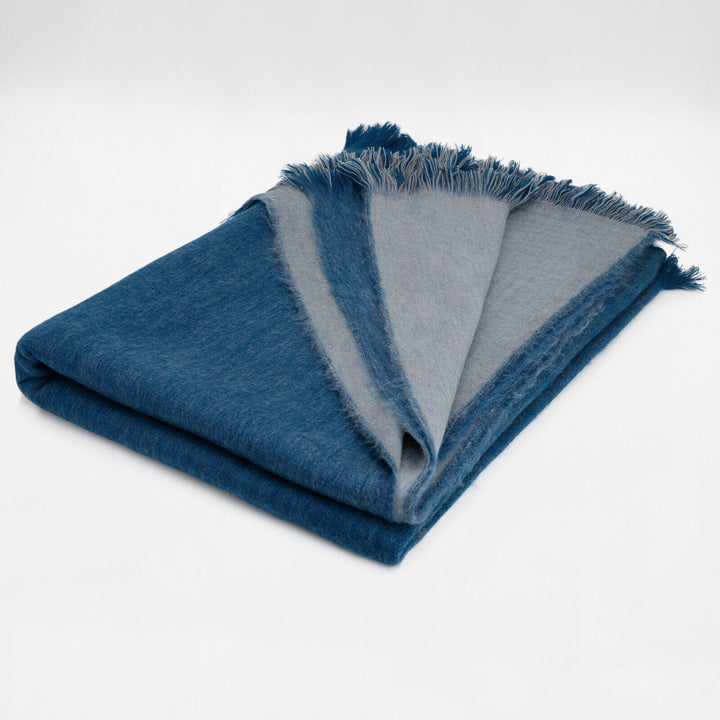 A folded, two-toned blue and gray blanket with fringed edges lies on a plain white background, showing its soft texture.