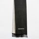 A dark grey woven scarf hangs vertically, featuring black fringe at the bottom and a white geometric design near the edge. The lighter underside is visible, set against a plain white background.