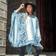 A person wearing a patterned blue poncho and black hat stands in a stone doorway, holding the brim of the hat while looking into the distance.