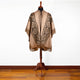 Brown poncho with geometric diamond patterns displayed on a wooden stand, featuring fringe details at the edges, situated on a polished wooden floor against a plain white background.