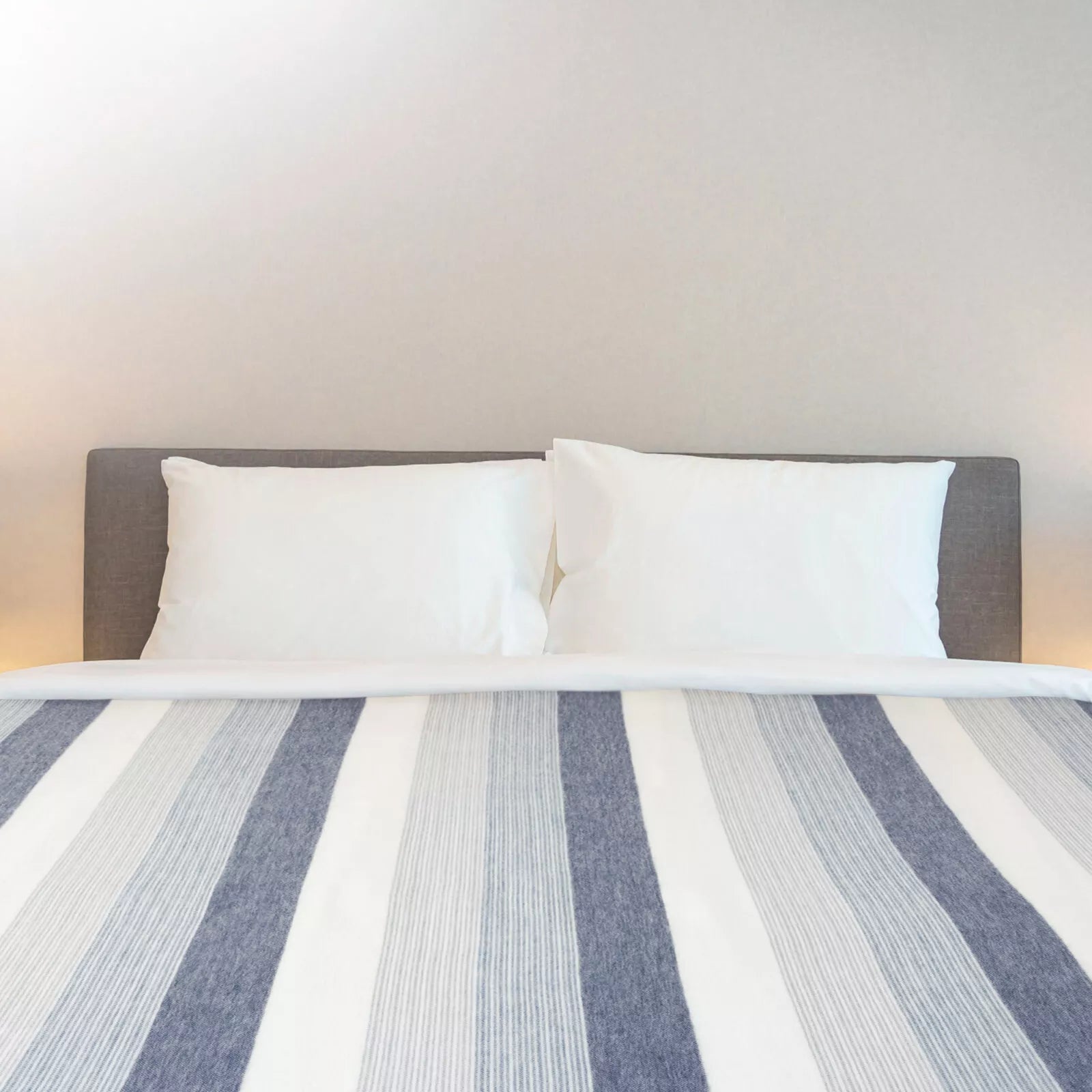 A neatly made bed displays two white pillows resting against a gray headboard, with a striped blue and white blanket covering most of the bed in a minimal, neutral room.